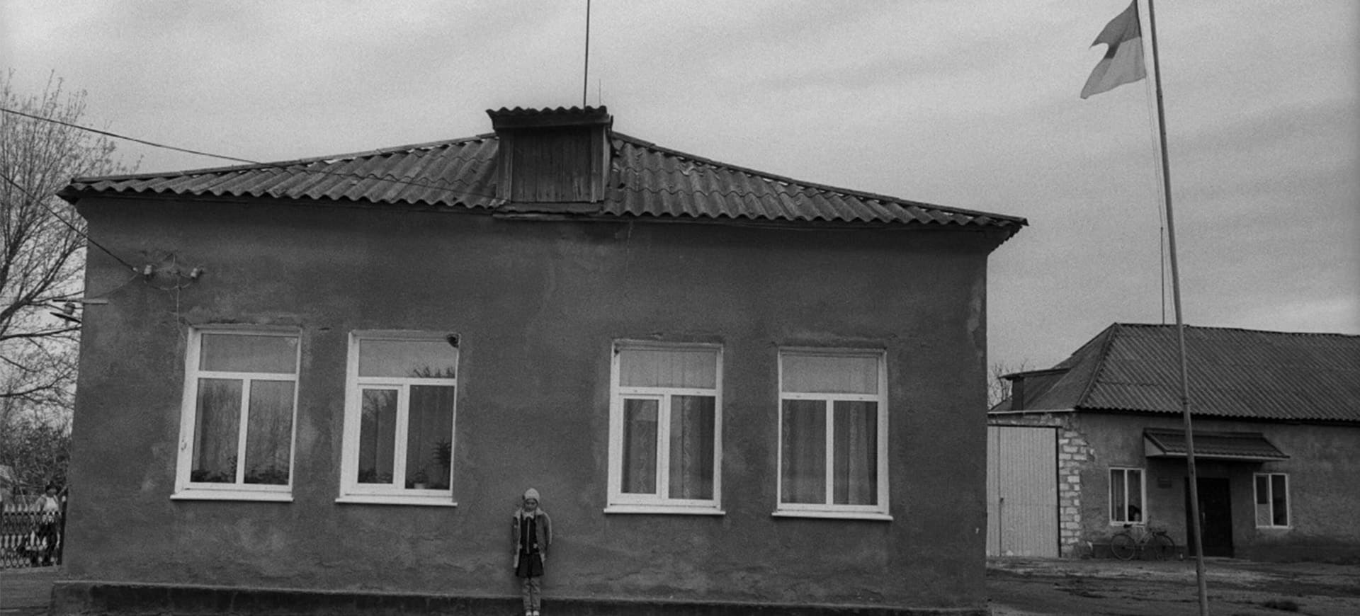 Black and White picture of a lone child warmly dressed, standing outside a soviet style orphanage building next to a ukrainian flag fluttering on a flagpole against a leaden sky