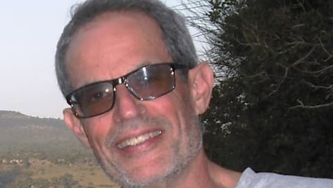 Close up of Loughborough University child development expert Professor Barry Bogin smiling at the camera. He stands in front of a green hill and tree. He's an older middle aged white man with short grey hair and stubble, dressed in a light grey t shirt and dark glasses. He looks happy and relaxed.