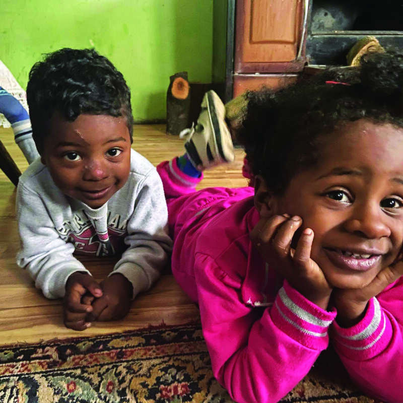 A young boy and girl lie on their front on the floor of their house, chins in hand looking up at the camera smiling