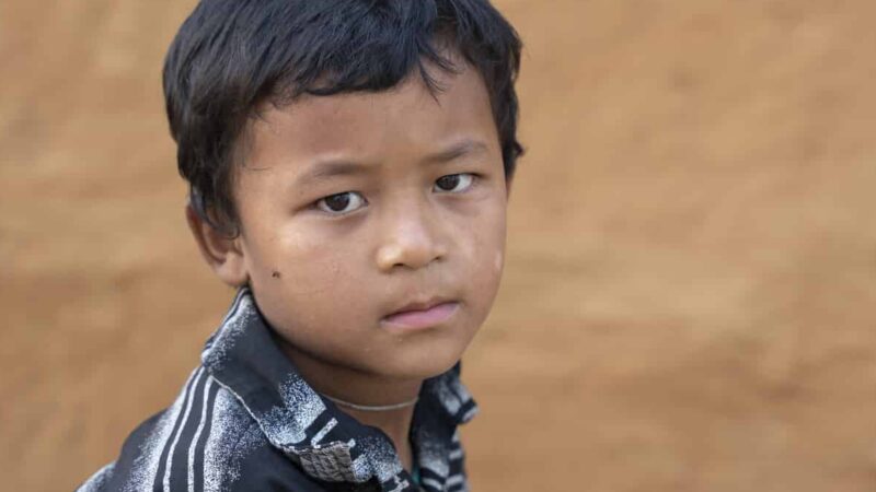 Young Nepali boy with short dark hair, in a striped polo shirt looks sadly sideways towards the camera, in front of brown earthen wall