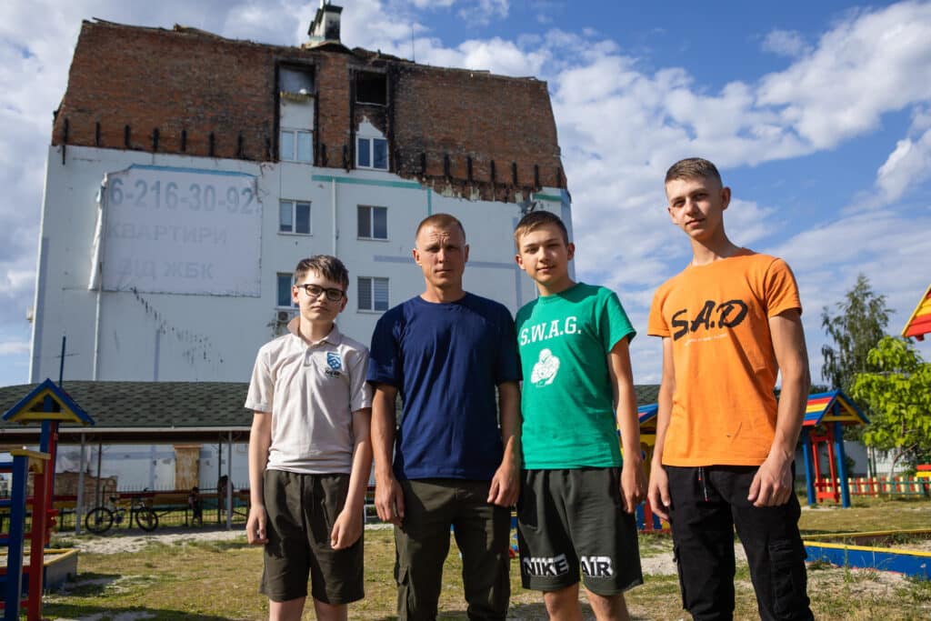Kyrylo, a man with blonde hair and a blue t shirt, stands in between his four nephews, all age between 11 and 19, in white, green and orange t shirts, in front of a large house in Ukraine. A fatherly figure