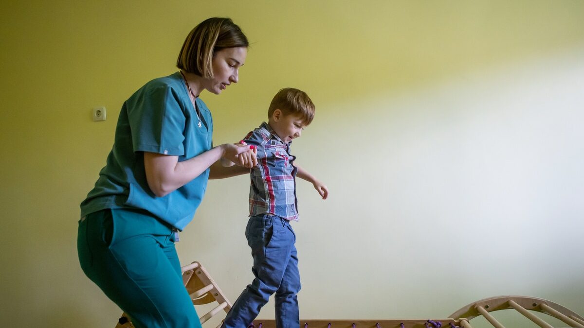 A support work wearing blue scrubs helps Nikita walk across a climbing frame.