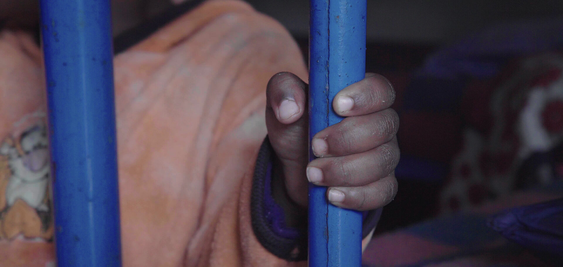 A child's hand holds a painted blue metal bar, locked in a dark orphanage