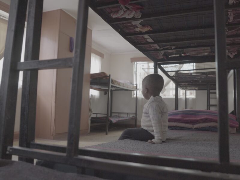 A young Kenyan boy sits alone on his bare metal bunk bed in an empty bleak dormitory