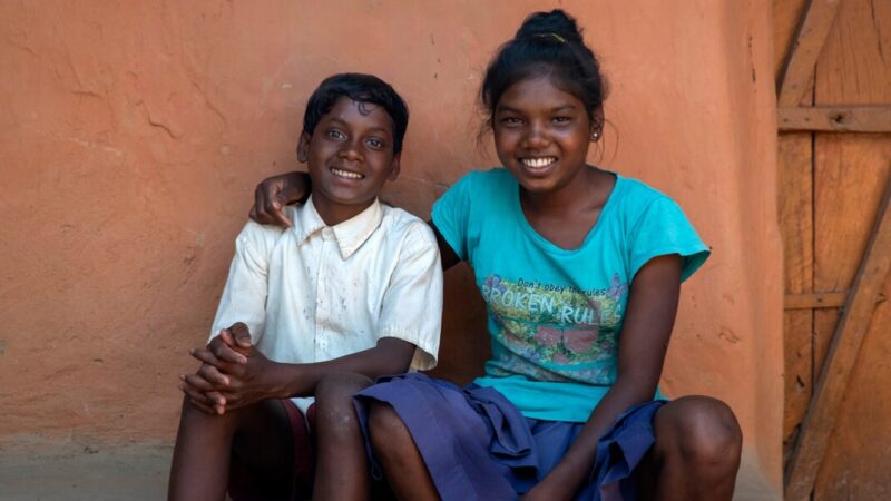 An Indian girl sits with her arm around her younger brother as they both smile at the camera
