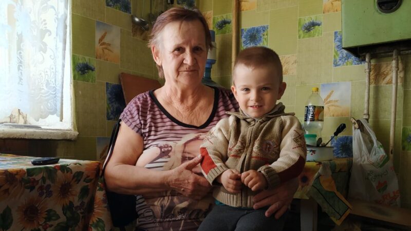 A young Ukrainian boy with short light brown hair, Oleksander, sitting on his grandmother's lap. They look confidently into the camera, in a pale yellow room.