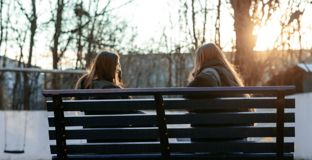 Two teenagers sit on a park bench, the sun shining behind them.