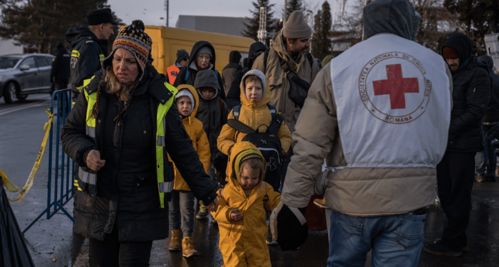 In celebration of World Refugee Day, this is a photo of support workers hold the hands of young children wearing bright yellow coats as they enter a border crossing zone.