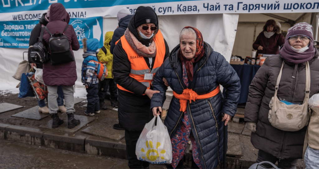 An elderly woman wearing a blue coat is helped by a support worker wearing an orange high-vis vest.