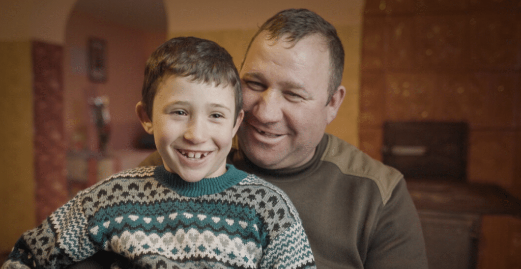 A father with short brown hair hugs his child, smiling. The child is a young boy, age seven, with brown hair and a green jumper.