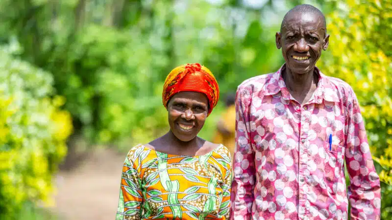 Two older Rwandan foster parents Masaro* and her husband Rugwiro* stand smiling in bright patterned shirts in front of greenery. They look proud and united.